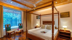 Una cama con dosel y cabecera de madera, dos mesitas de noche con lámparas, una pequeña mesa redonda con una silla y vistas al exterior a través de grandes ventanales.