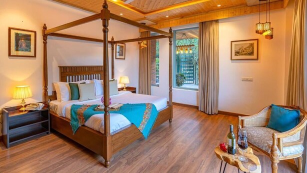 Una cama con dosel y cabecera de madera, una mesita de noche con lámpara, una silla de mimbre y una ventana con cortinas.