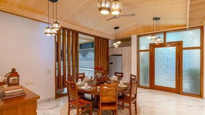 Comedor con una mesa redonda dispuesta para seis personas, rodeada de sillas de madera. Hay lámparas colgantes sobre la mesa, y se ve un ventilador de techo. La habitación tiene paredes de madera y un piso de baldosas.