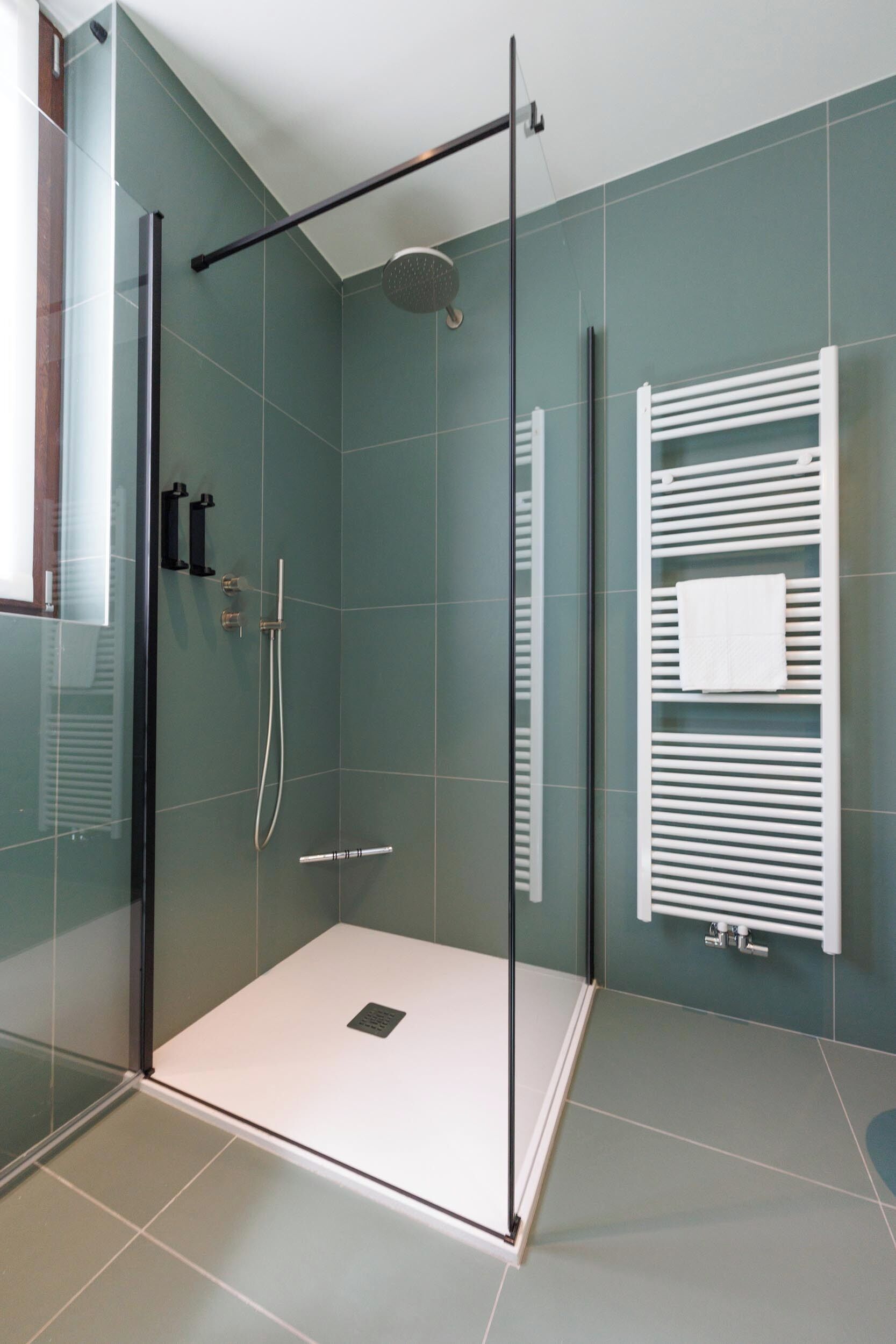 Un área de ducha moderna con puerta de vidrio, cabezal de ducha manual y un toallero blanco.