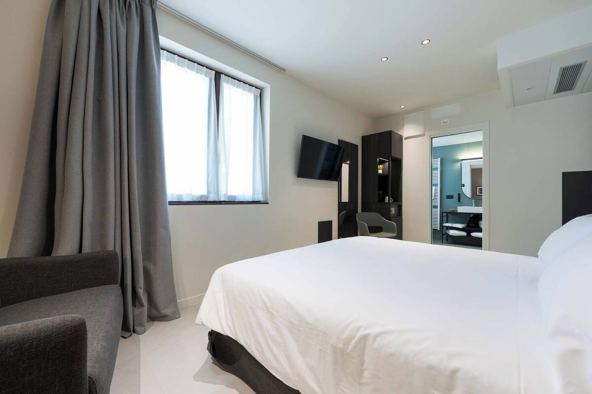 Una habitación de hotel con una cama grande, un televisor montado en la pared, un escritorio con una silla y un baño visible a través de una puerta abierta.