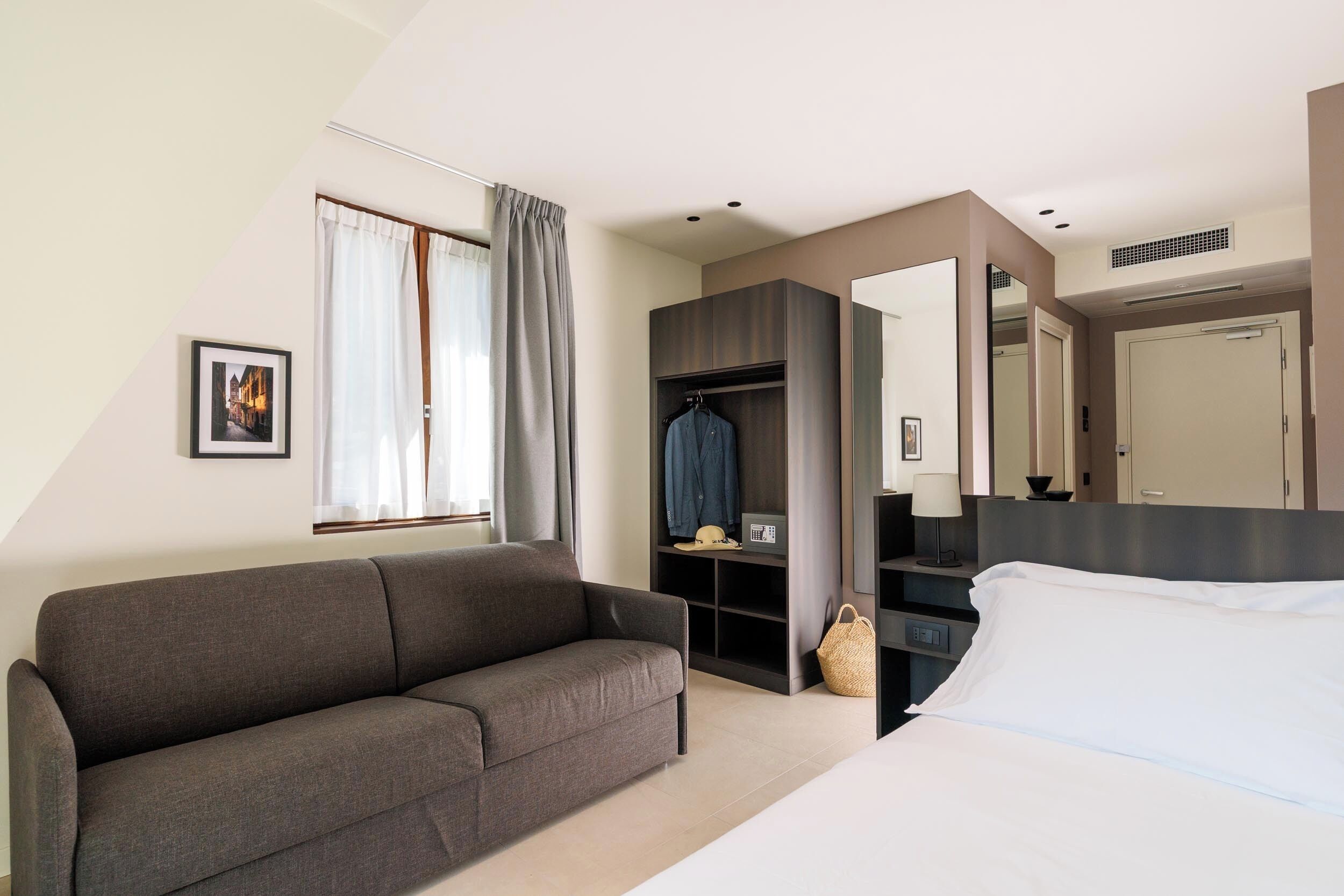 A modern hotel room with a gray sofa, a bed with white linens, a built-in wardrobe, a large mirror, and a picture on the wall.