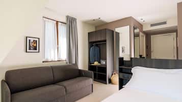 A modern hotel room with a gray sofa, a bed with white linens, a built-in wardrobe, a large mirror, and a picture on the wall.