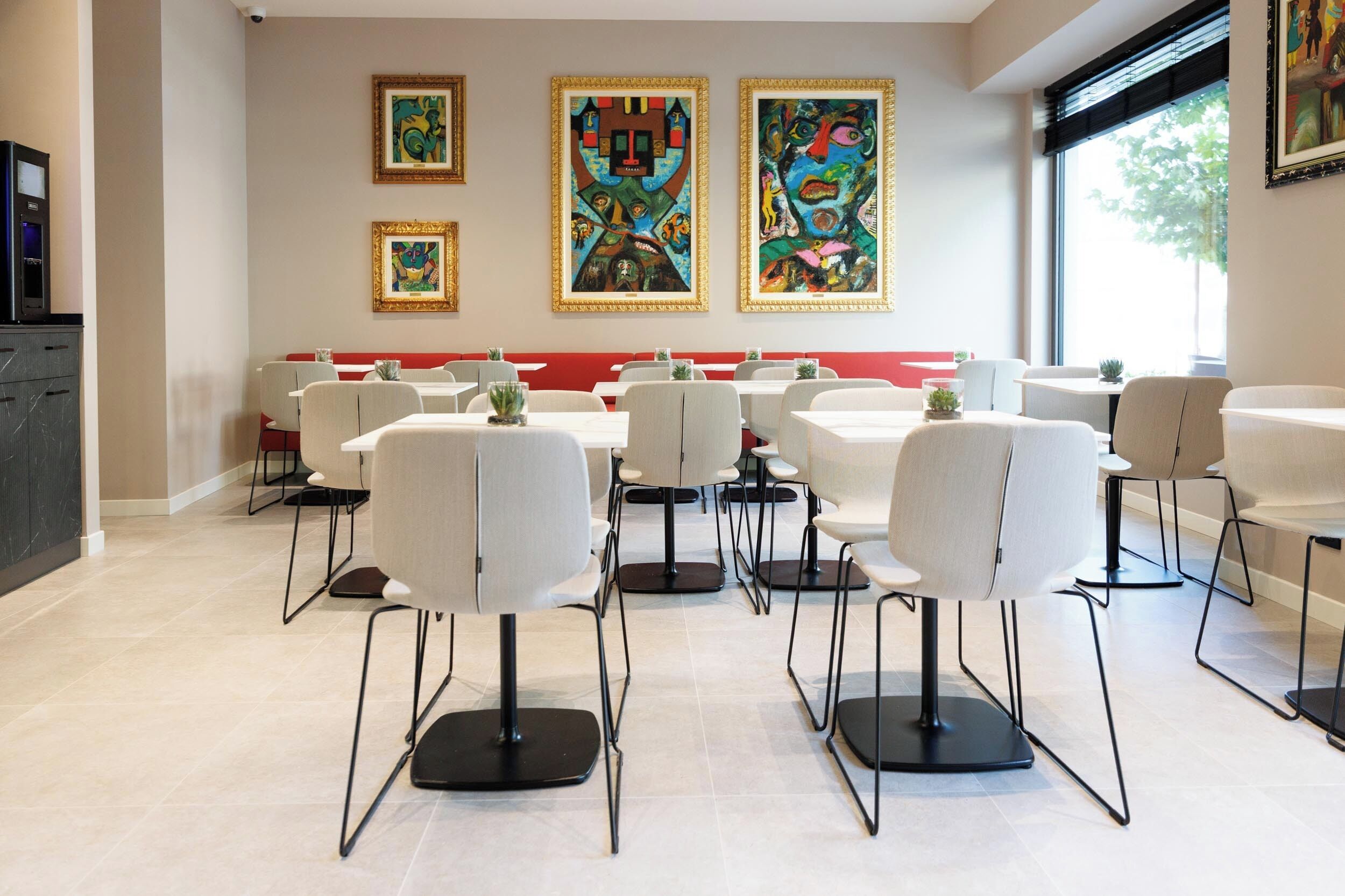 A modern dining area with white chairs and tables, a red bench, and colorful abstract paintings on the wall.