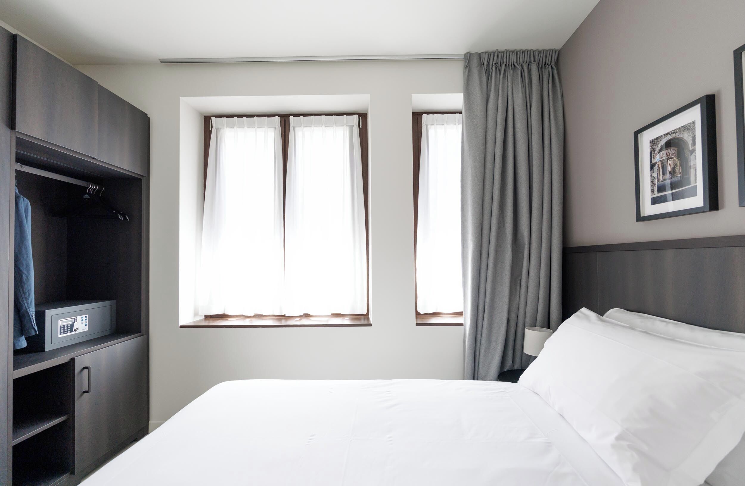 Habitación de hotel con cama, armario, dos ventanas con cortinas y un cuadro en la pared.