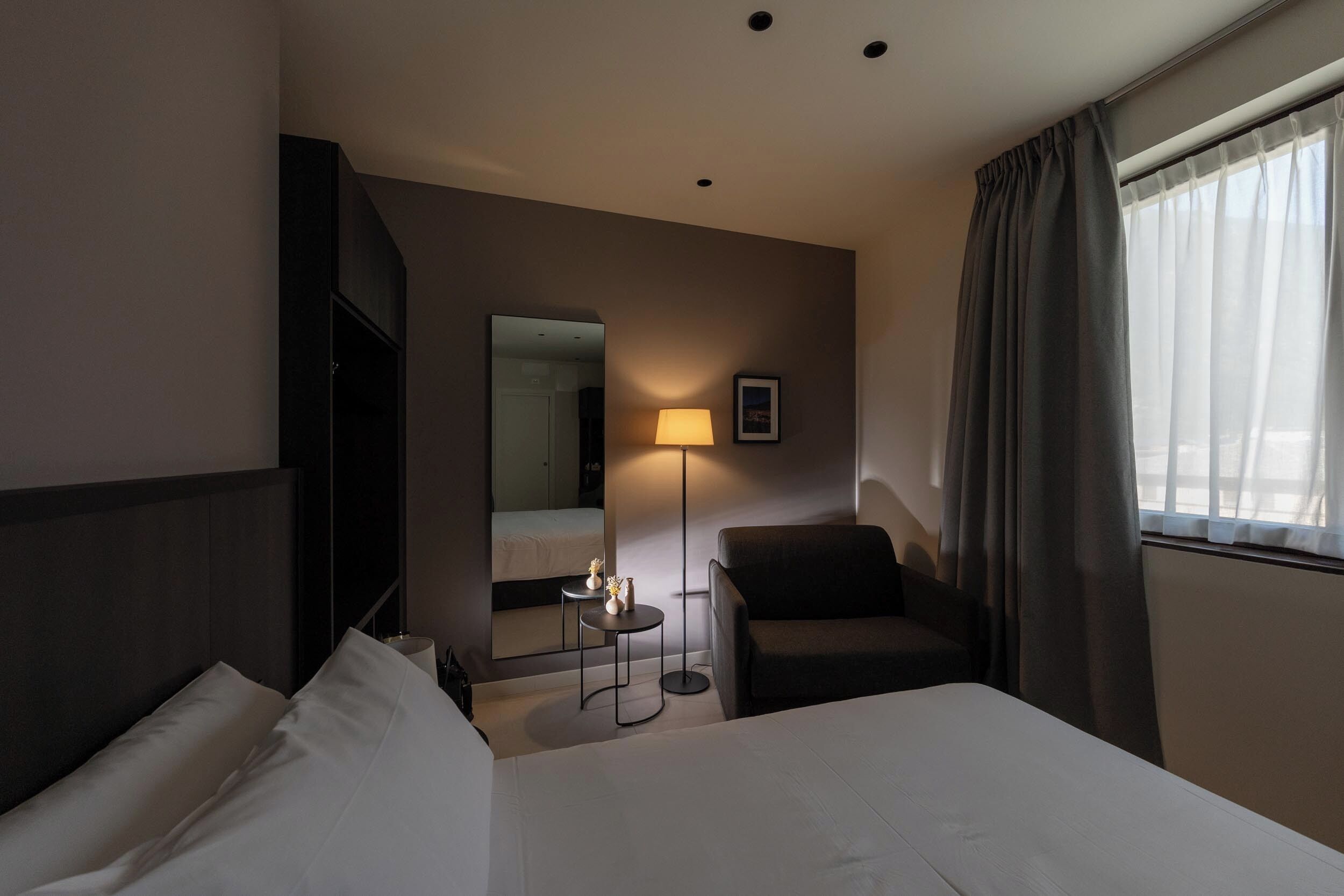 A hotel room with a bed, a chair, a desk, a lamp, a mirror, and a window with curtains.