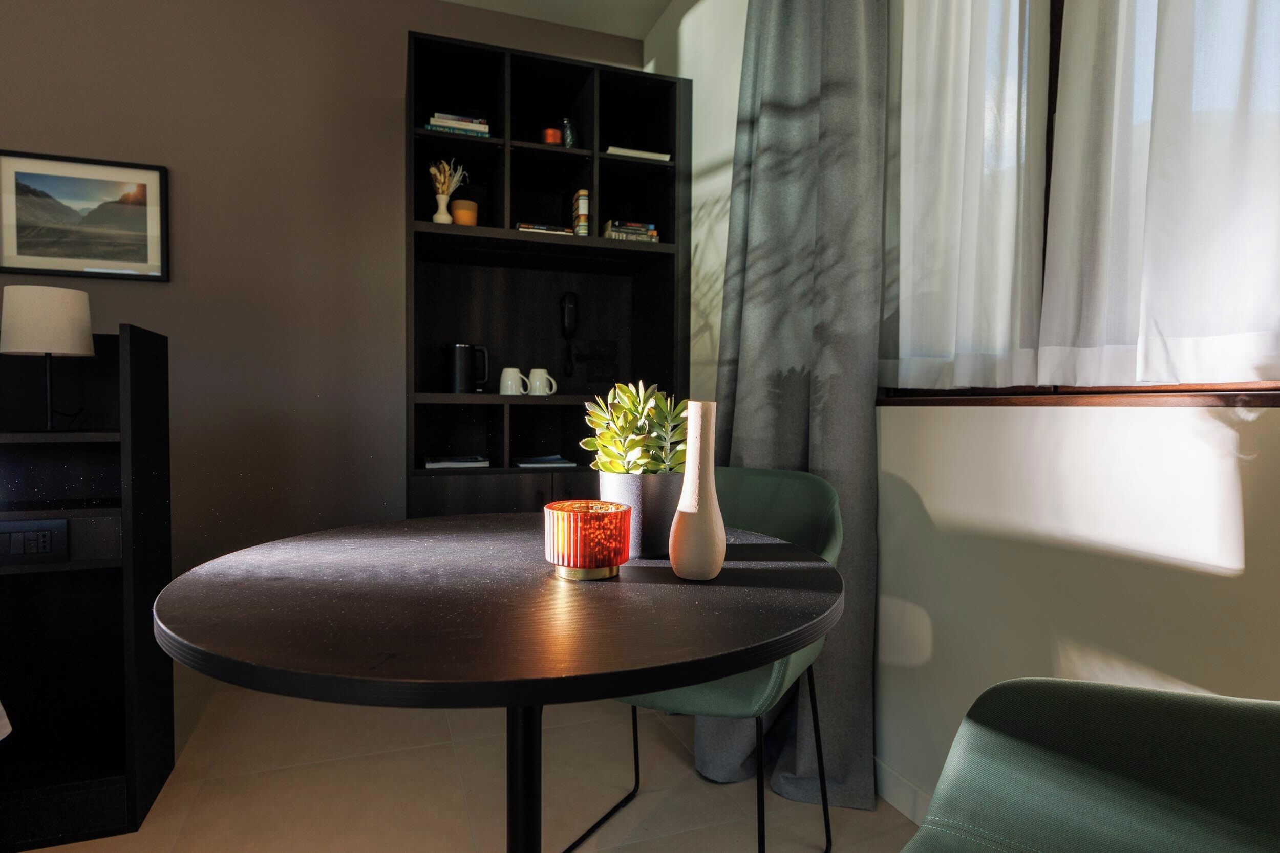 Une table ronde avec un vase et une plante, une bibliothèque et une chaise verte dans une pièce dotée d’une fenêtre et de rideaux.
