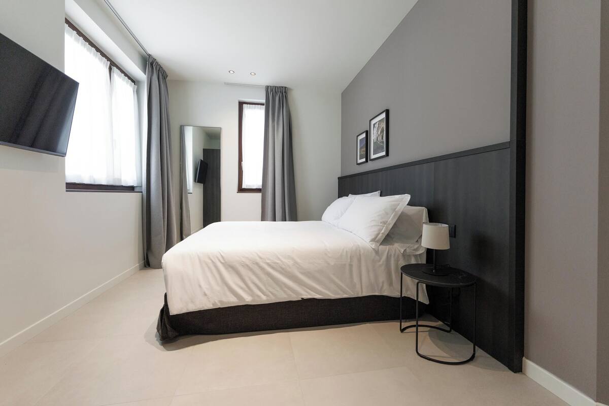 Una habitación de hotel moderna con una cama grande, una mesita de noche con lámpara, un televisor de pantalla plana en la pared y una ventana con cortinas.