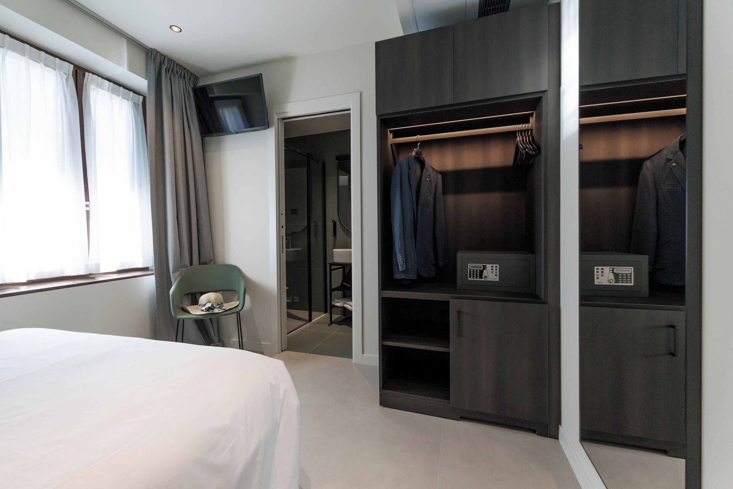 A hotel room with a bed, a green chair, a walk-in closet with hanging clothes, and a TV mounted on the wall.