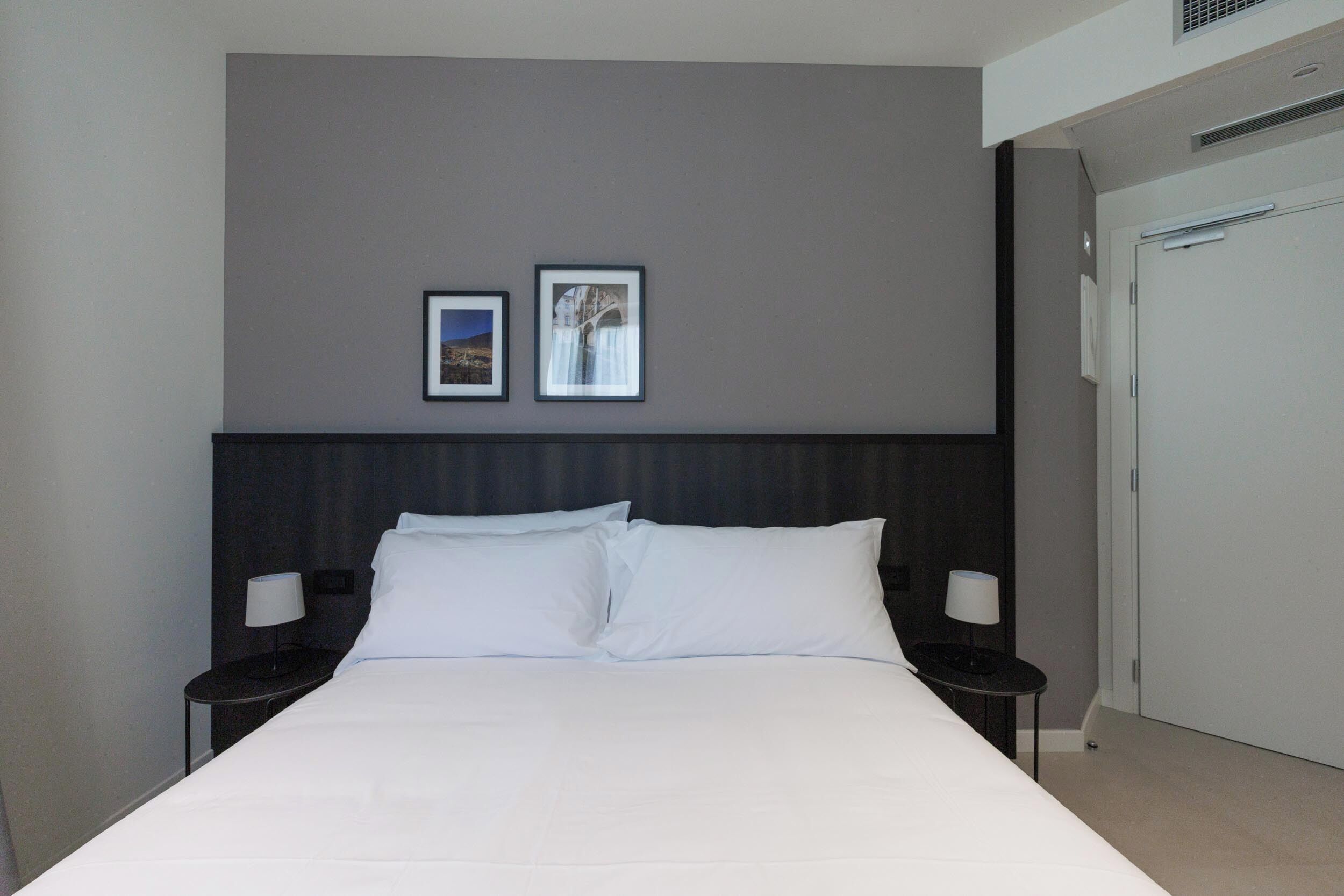 A hotel room with a large bed, two bedside tables with lamps, a dark headboard, and two framed pictures on the wall.