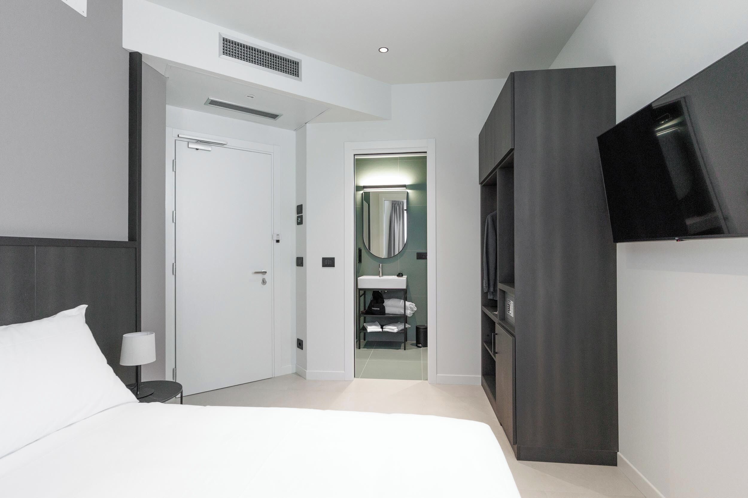 A modern hotel room with a bed, a TV, a wardrobe, and a bathroom visible through an open door.