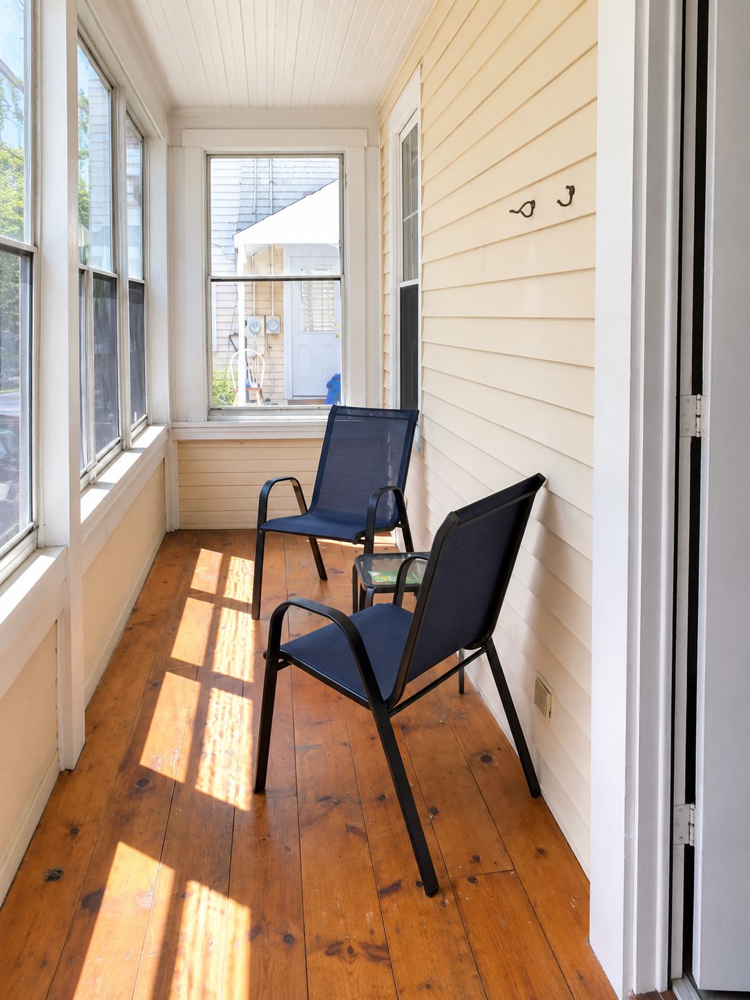 Habitación iluminada por el sol con suelo de madera, dos sillas, una mesita y una ventana con persianas.