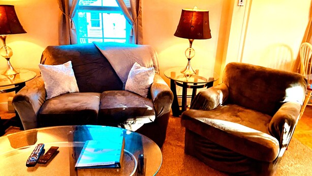 A living room with a sofa, armchair, and glass coffee table. There are two lamps on side tables and a window with curtains. - Portland 1-bedroom with enclosed porch & parking (Portland)