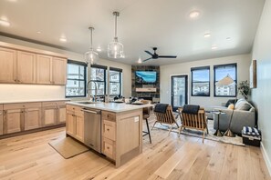 A modern kitchen with wooden cabinets, a central island, pendant lights, and a dining area with a sofa and chairs. - Flying Moose At Helling Circle 101 3 Bedroom Condo (Heber City)