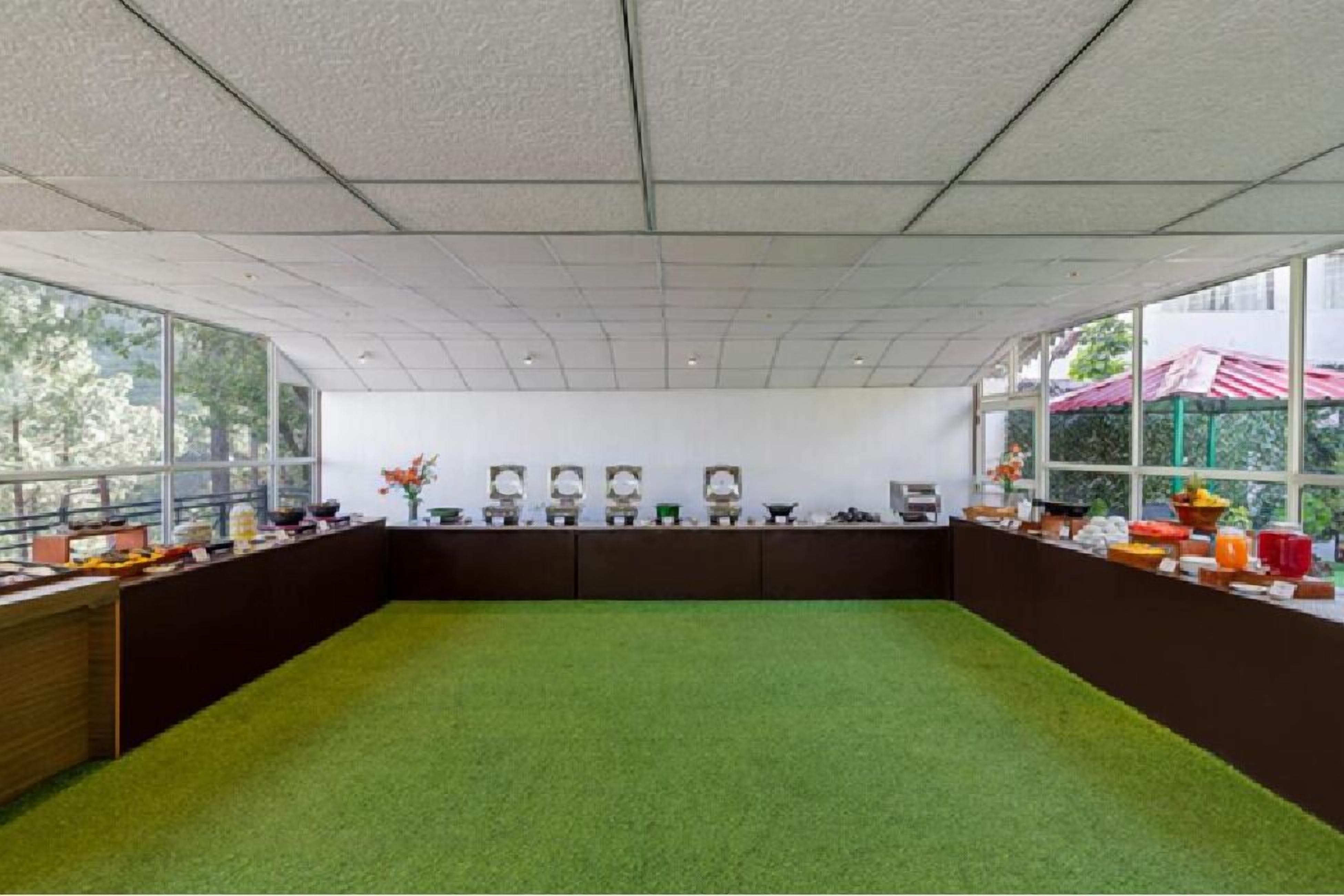 Une pièce avec un tapis vert, une longue table sur laquelle se trouvent divers objets, et de grandes fenêtres.