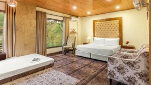 A room with a wooden ceiling, a bed with a tufted headboard, a chair, a small table with a lamp, and a window with curtains. - Maple Resort Chail (Chail)