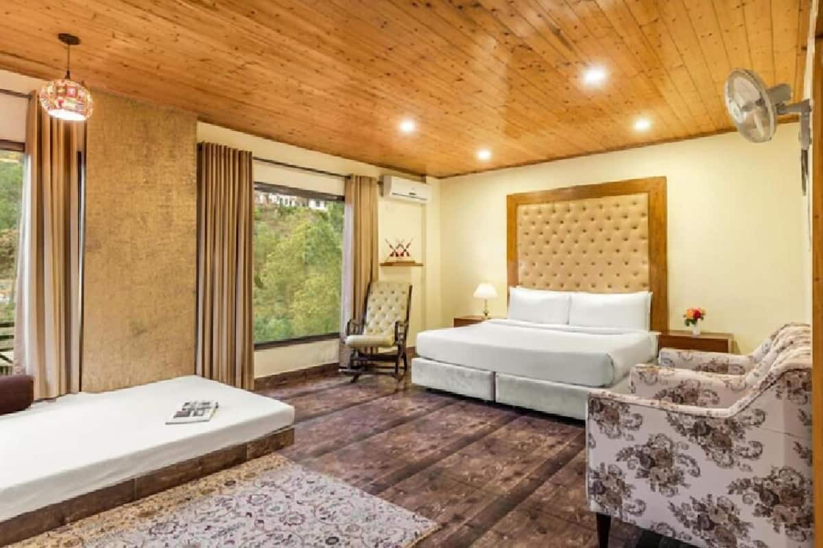 Une pièce avec un plafond en bois, un lit doté d’une tête de lit capitonnée, une chaise, une petite table avec une lampe et une fenêtre avec des rideaux.