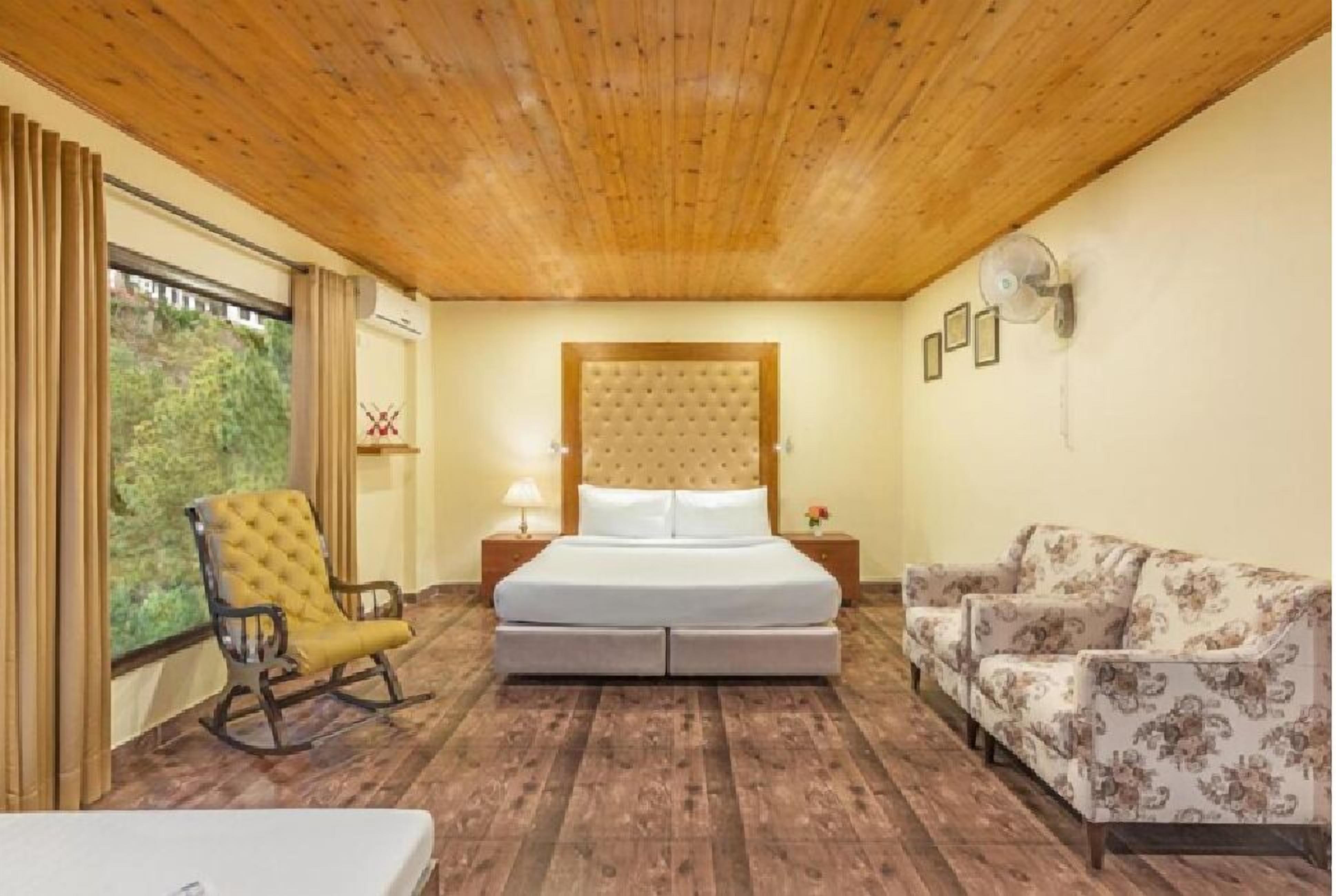 A bedroom with a wooden ceiling, a bed with a tufted headboard, a rocking chair, a sofa, and a nightstand with a lamp.
