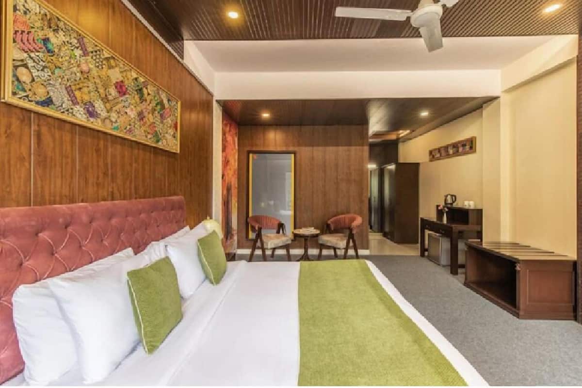 Une chambre d’hôtel avec un grand lit, un plaid vert, une tête de lit rembourrée en tissu rouge, un ventilateur de plafond et un mur en bois orné d’une œuvre d’art colorée.
