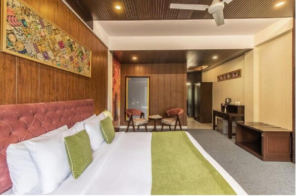 A hotel room with a large bed, a green throw blanket, a red upholstered headboard, a ceiling fan, and a wooden wall with a colorful artwork. - Maple Resort Chail (Chail)