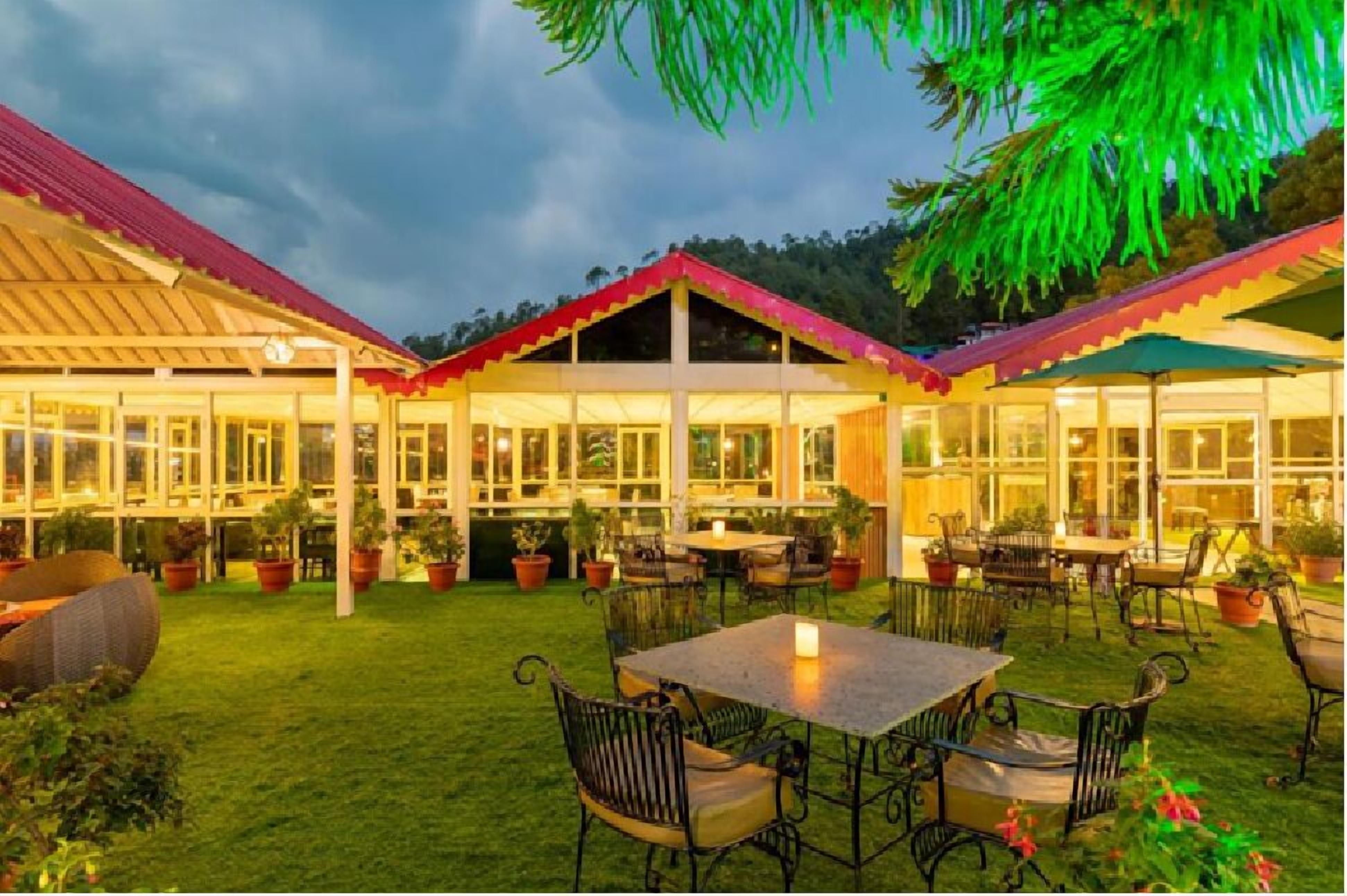 Un área de comedor exterior bien iluminada con mesas y sillas, rodeada de plantas en macetas y un edificio con techo rojo.