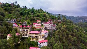 A cluster of multi-story buildings with red roofs nestled among dense green trees on a hillside. - Maple Resort Chail (Chail)