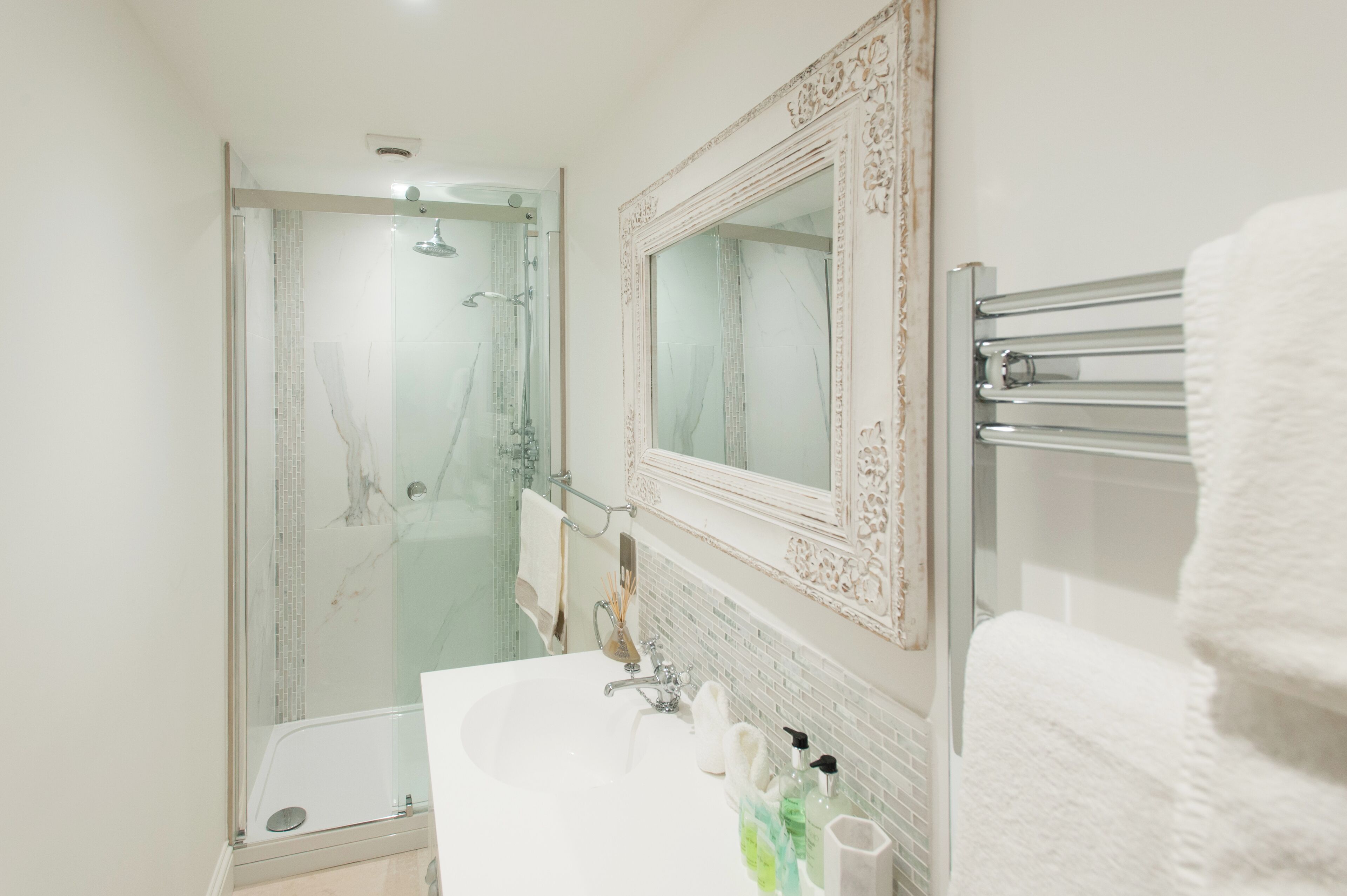 Un baño con ducha de vidrio, un lavamanos blanco con espejo y un perchero con una toalla blanca.