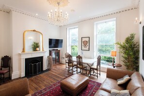 A room with a fireplace, a television, a dining table with chairs, a sofa, and a large window with a view of greenery. - 21 Montpellier Terrace (Cheltenham)