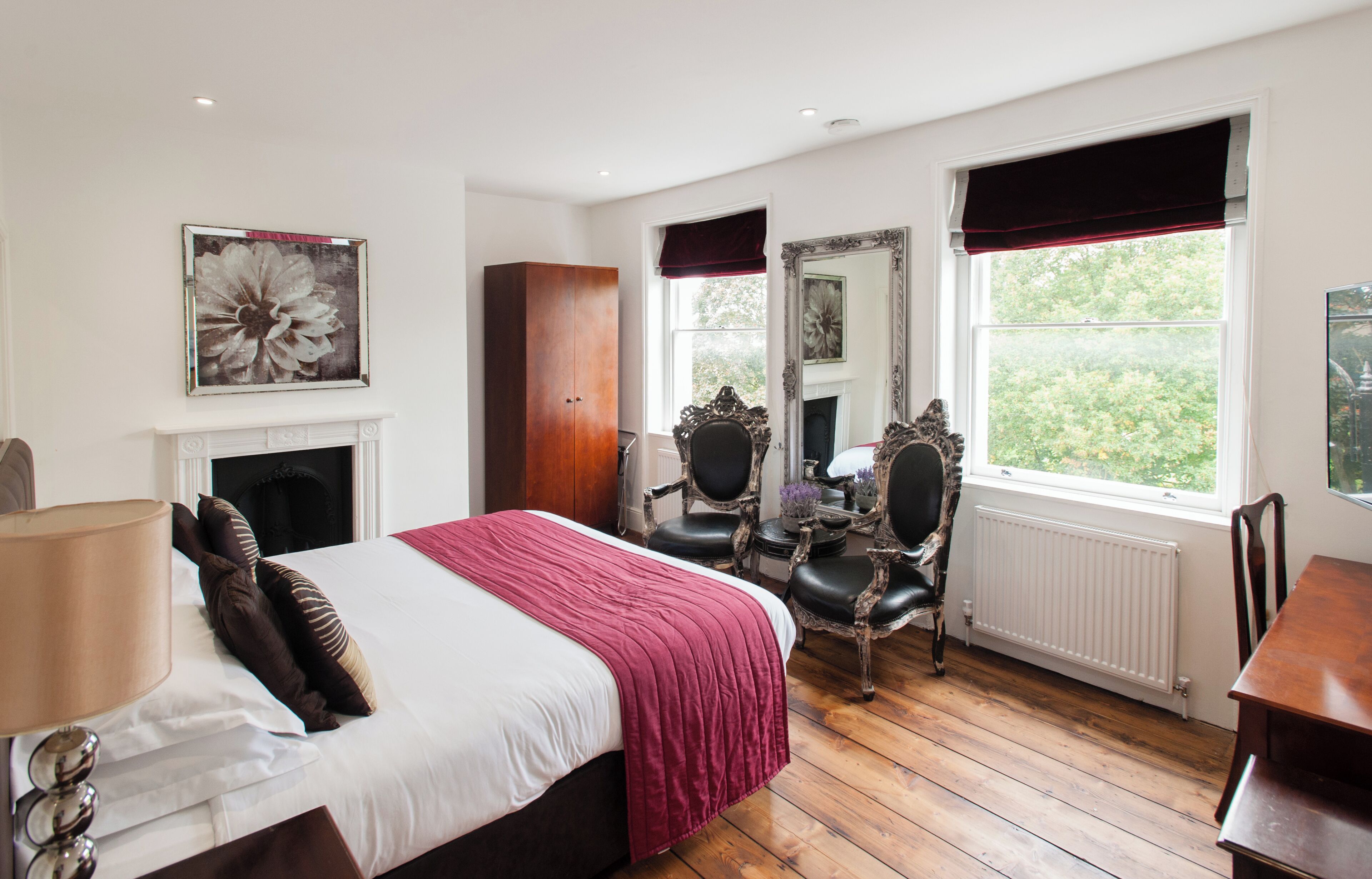 A bedroom with a bed, a fireplace, a mirror, and a window with a view of greenery.