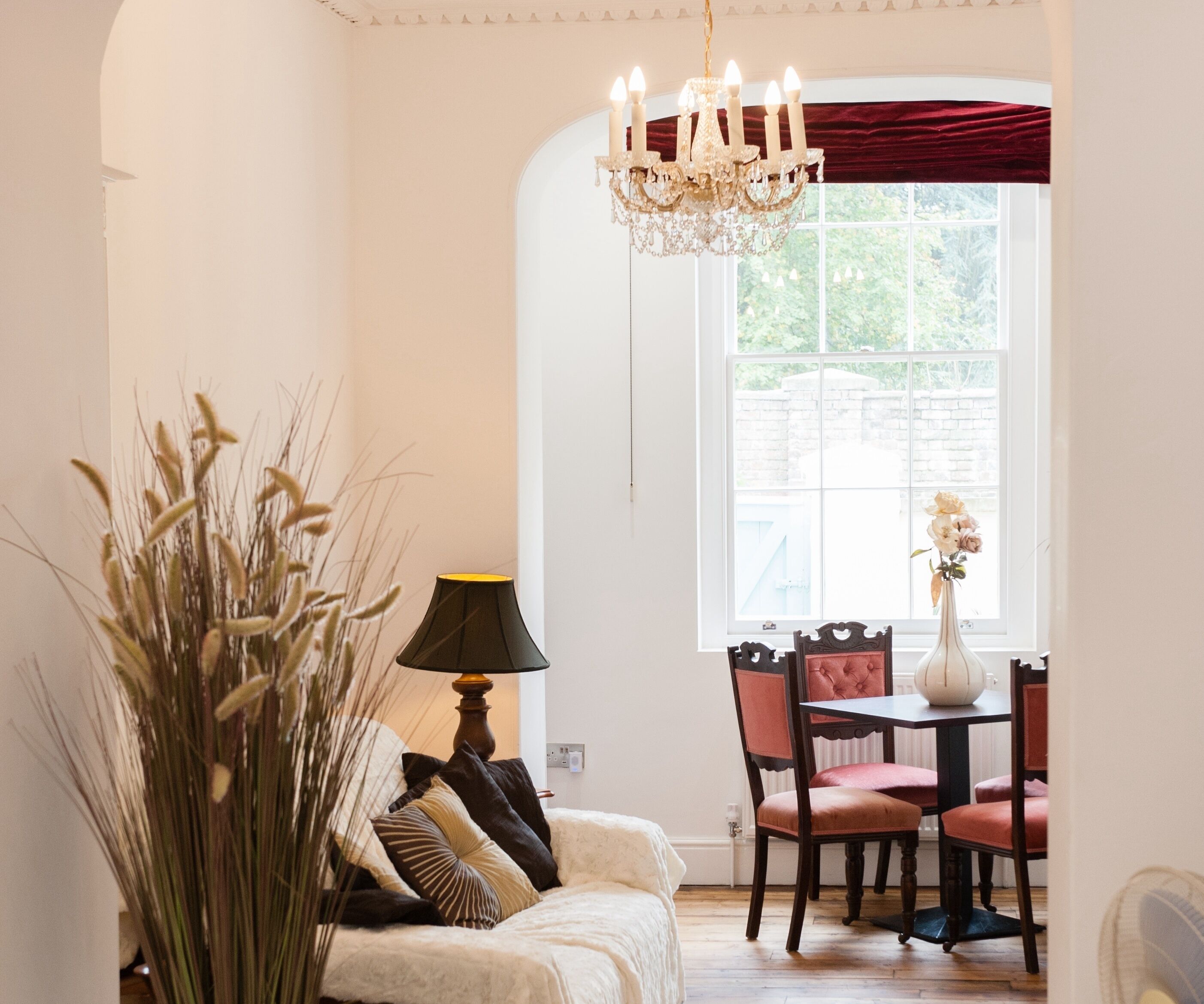 A cozy room with a white sofa, a dining table with chairs, a chandelier, and a window with a view of greenery.