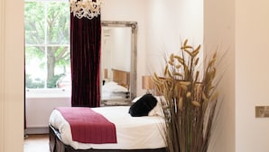 A bedroom with a large mirror, a bed with a white and red bedspread, a dark headboard, and a window with a view of trees. - 21 Montpellier Terrace (Cheltenham)