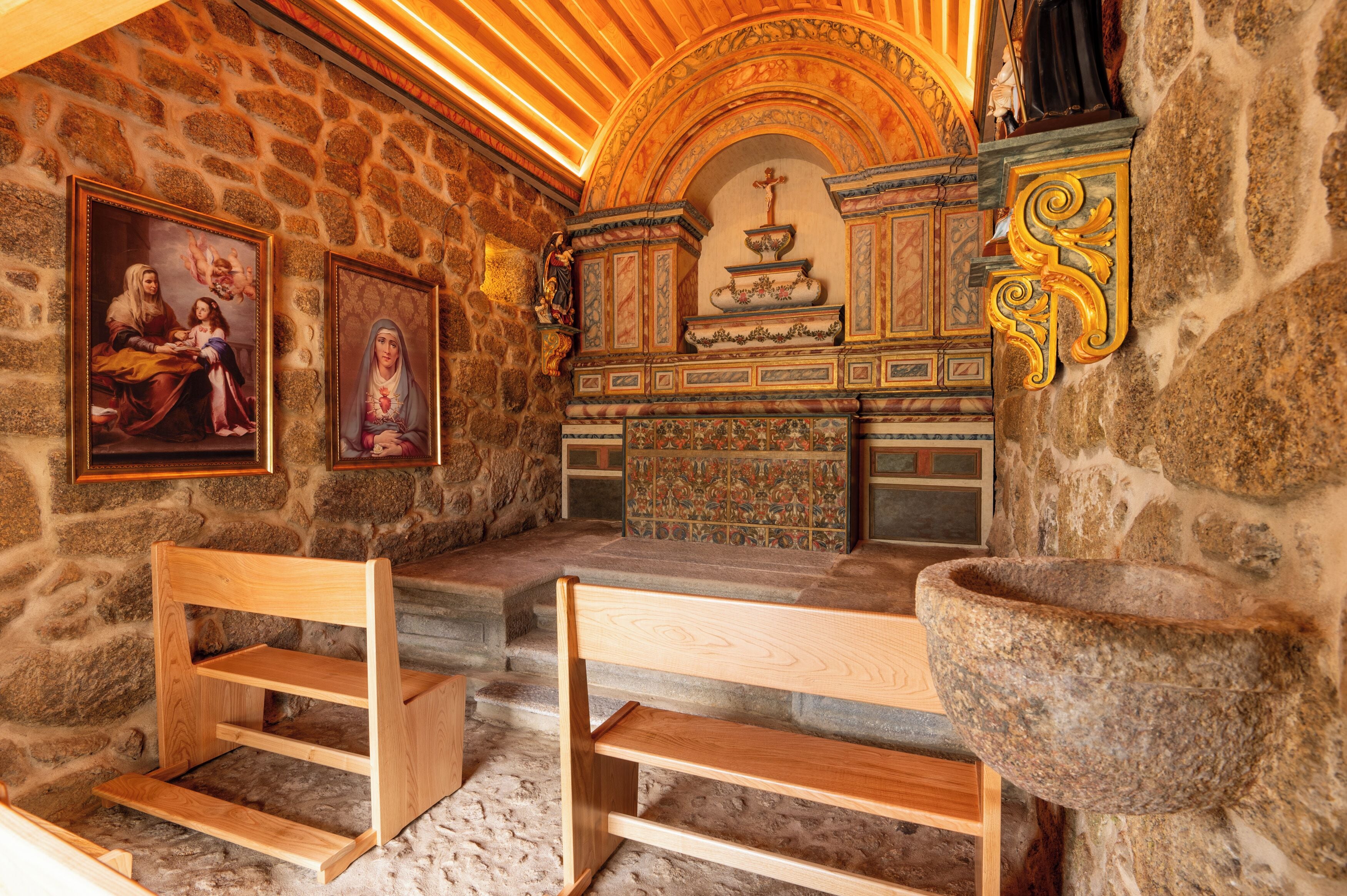 Una capilla de paredes de piedra con bancos de madera, una pila de piedra para bautismos y pinturas religiosas.