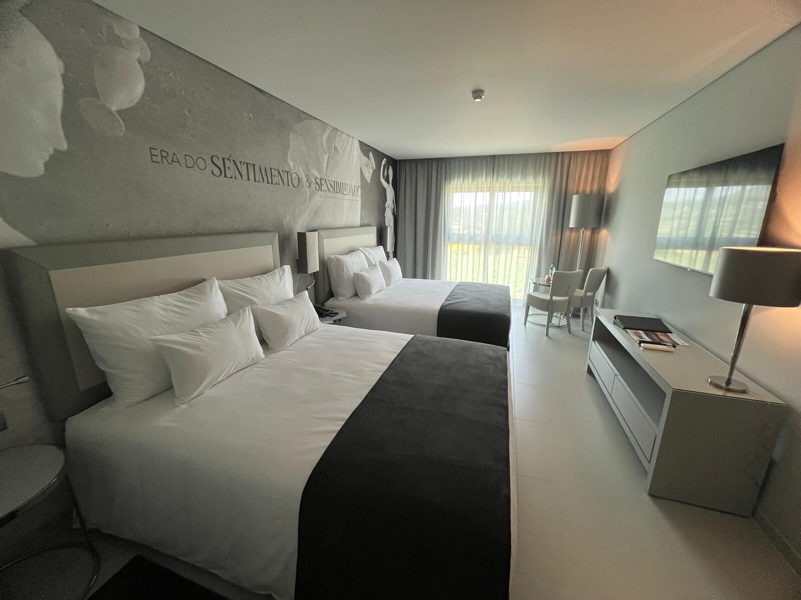 A hotel room with two beds, a desk, a chair, and a large window with curtains.