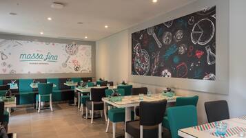 Interior de restaurante con sillas verde azulado y negras, mesas blancas y una pared de pizarra con ilustraciones de comida.
