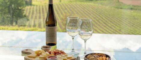 A table with a bottle of wine, two glasses, a cheese board, and a bowl of snacks on a balcony with a vineyard view.