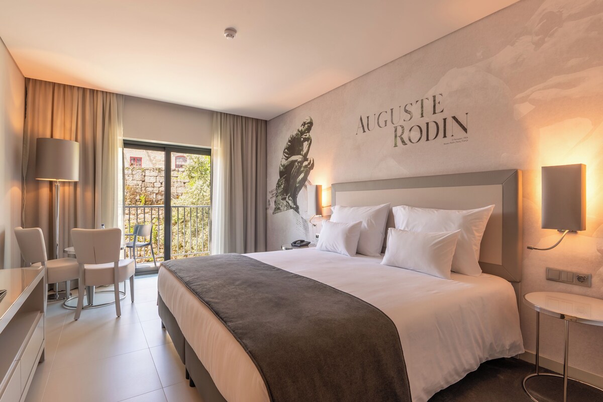 Een moderne hotelkamer met een groot bed, een bureau en een stoel. Er is een raam met gordijnen, een wandlamp en een muur met een naambordje.