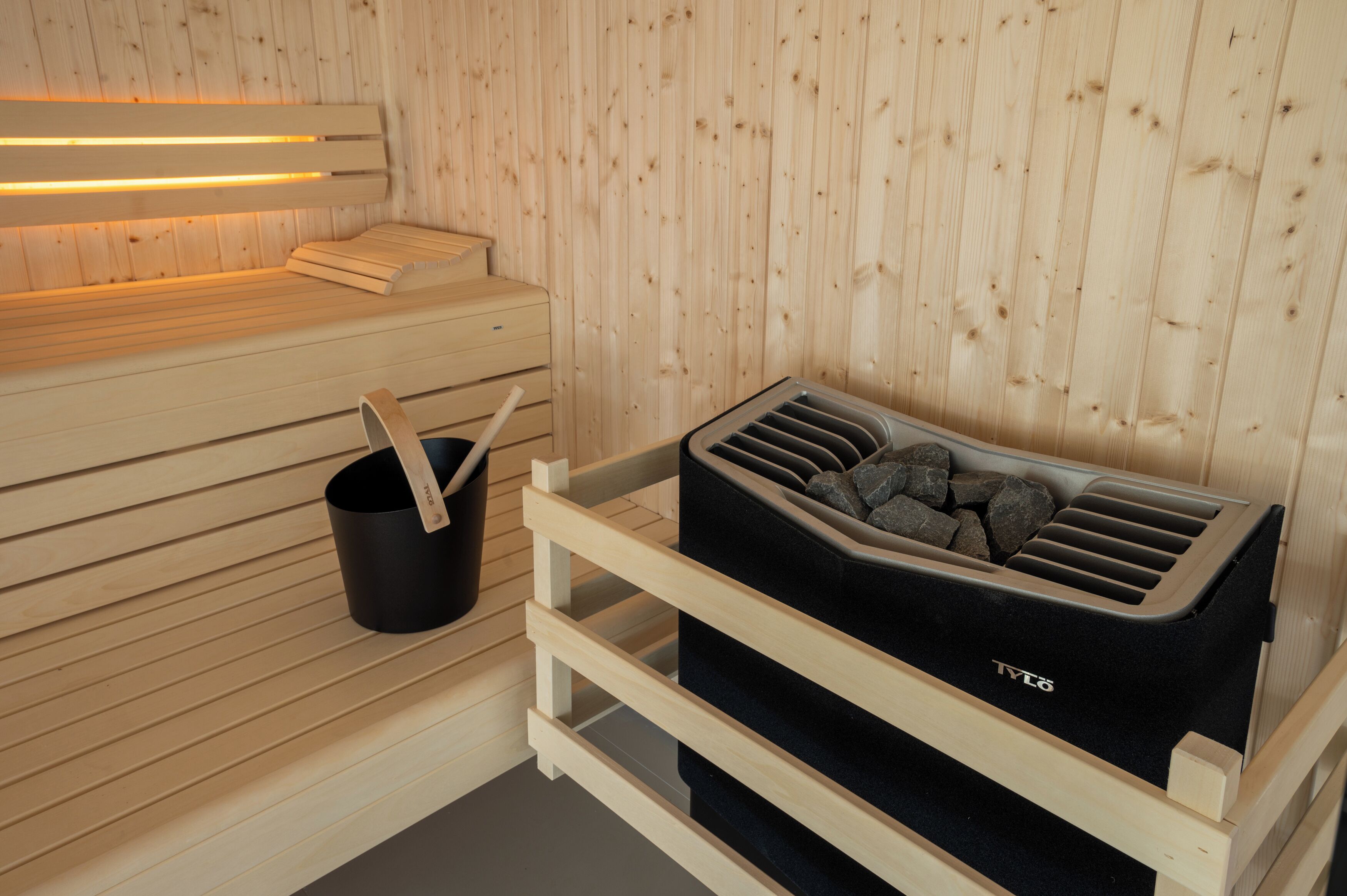 A sauna with wooden benches, a black container with rocks, and a black bucket with a wooden handle.