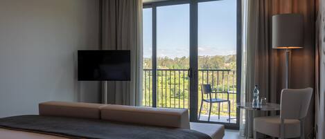 A modern hotel room with a large bed, a flat-screen TV, a balcony with a table and chairs, and a view of the outdoors.