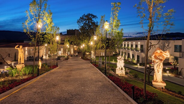 Un camino empedrado bordeado por árboles y estatuas, que lleva a un edificio de fachada de piedra.