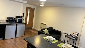 A compact kitchen with a microwave, toaster, and coffee maker. There is a dining area with a table set for two, including plates, cutlery, and a glass. A black sofa and a small side table with a picture frame are also visible. - 6 Bed Apartment @ Astor House by Pureserviced (Plymouth)