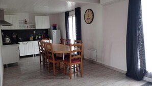 Un coin repas avec une table et des chaises en bois, une cuisine à l’arrière-plan et une horloge murale.