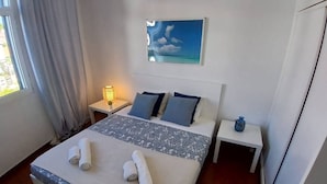 A hotel room with a bed, two bedside tables, a nightstand, a lamp, a painting, and a window with curtains.