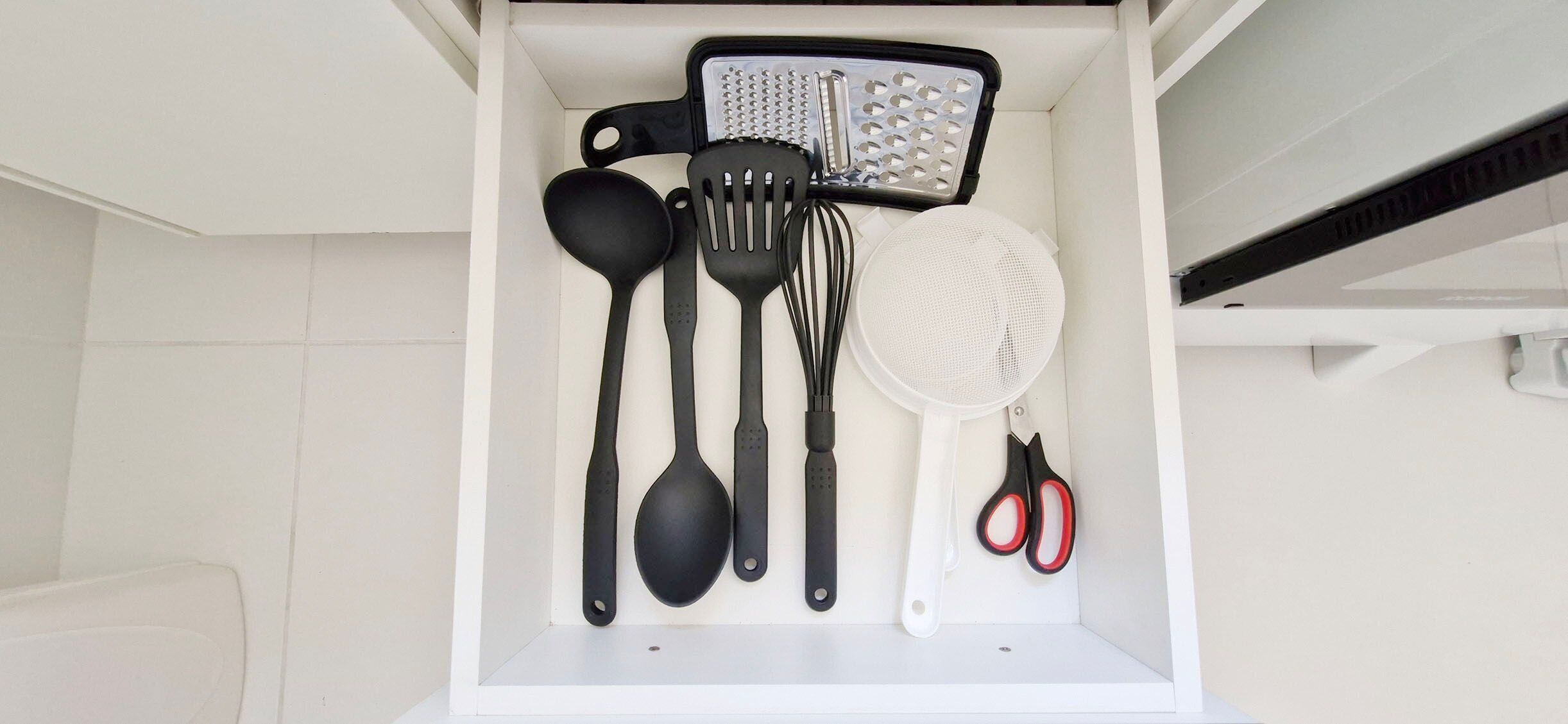 Un cajón de cocina con varios utensilios, como un rallador, espátula, cuchara, batidor, colador y tijeras.