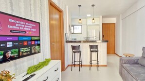 A modern living room with a grey sofa, a TV mounted on the wall displaying a streaming service, and a kitchen area with a bar counter and two stools. - COGRA0102 - Edifício Graça Prime (Salvador)
