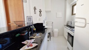 A compact kitchen with a black countertop, stainless steel sink, white cabinets, a gas stove, and a white refrigerator. - COGRA0102 - Edifício Graça Prime (Salvador)