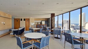A modern hotel lobby with seating areas, a kitchen, and large windows offering a city view. - COGRA0102 - Edifício Graça Prime (Salvador)