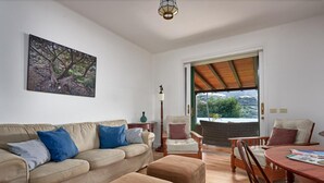 Ein Wohnzimmer mit einem beigen Sofa, zwei Poufs, einem Holzesstisch und Stühlen. An der Wand hängt ein Gemälde und das Fenster bietet Blick auf Grünflächen.