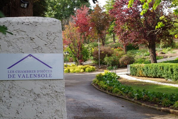 Schild mit der Aufschrift 'Les Chambres d'hôtes de Valensole', Blick auf einen angelegten Garten und eine Straße.