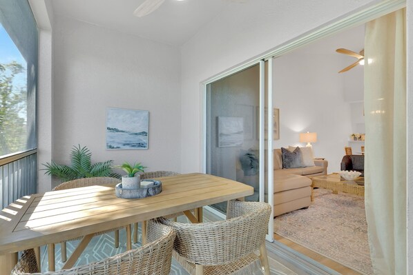 A sunlit room with a wooden table, wicker chairs, and a view through a glass door to a living area with a sofa and lamp.
