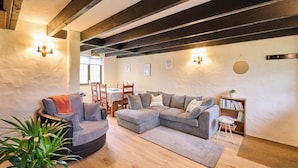 A cozy living room with a sectional sofa, a round chair, and a bookshelf. There is a dining table with chairs in the background. - Host & Stay | Providence Cottage (Woolacombe)